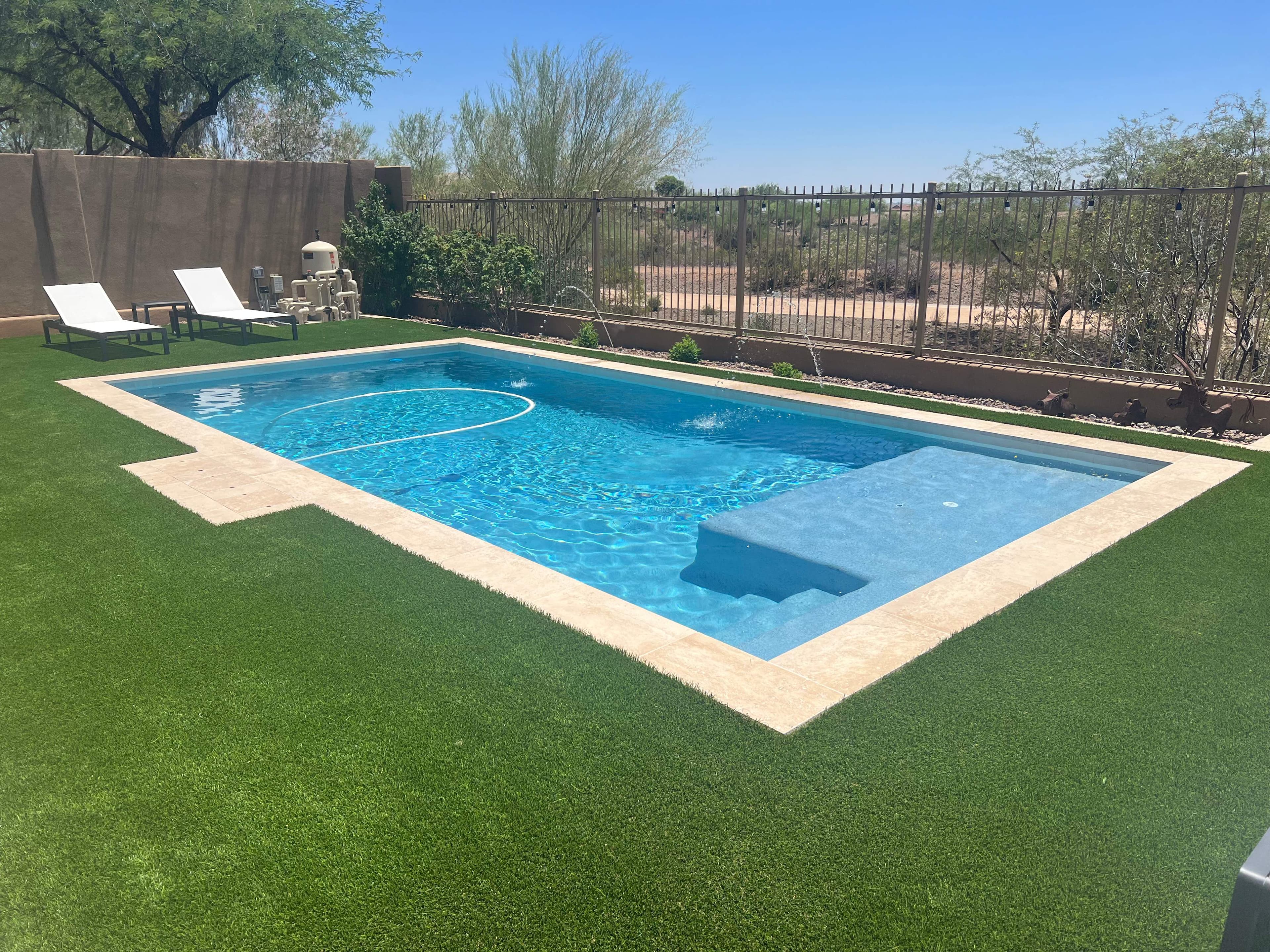 Rectangular blue pool with a shallow Baja shelf and steps, travertine coping, artificial turf, and desert landscaping beyond a view fence