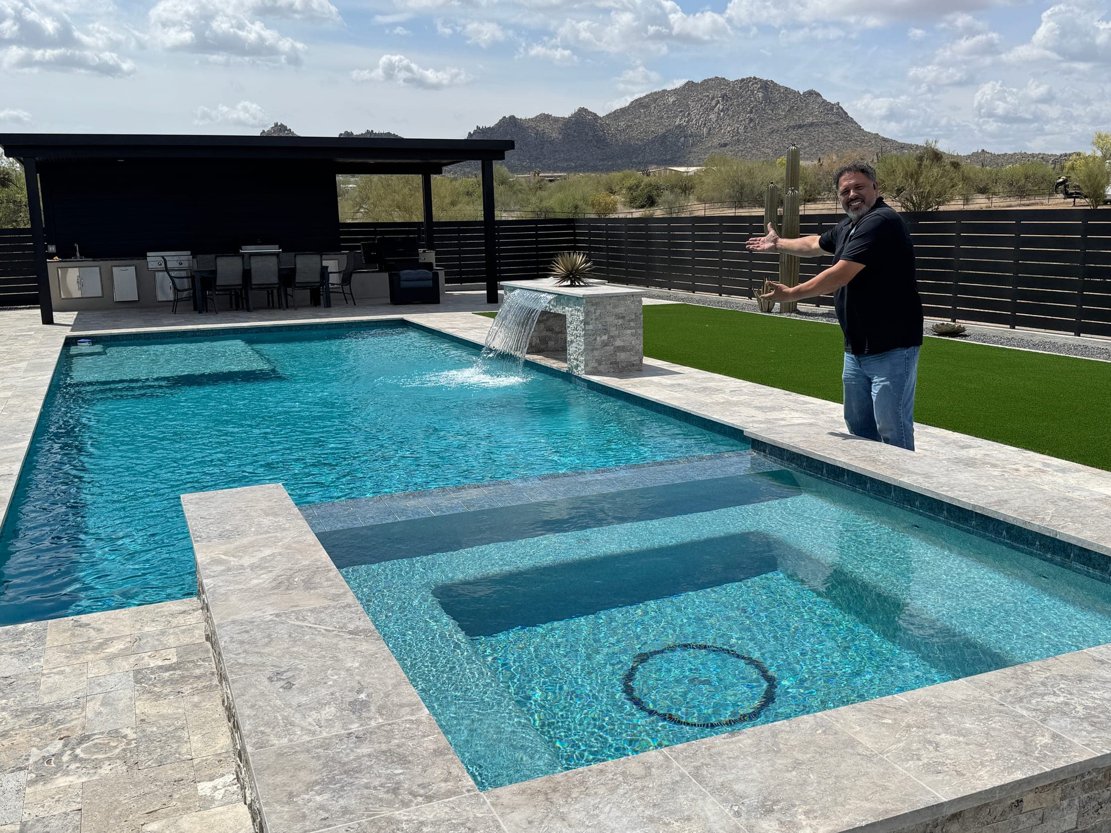 Completed geometric pool and raised spa with a sheer descent, travertine deck, turf, and a covered outdoor kitchen against desert mountains