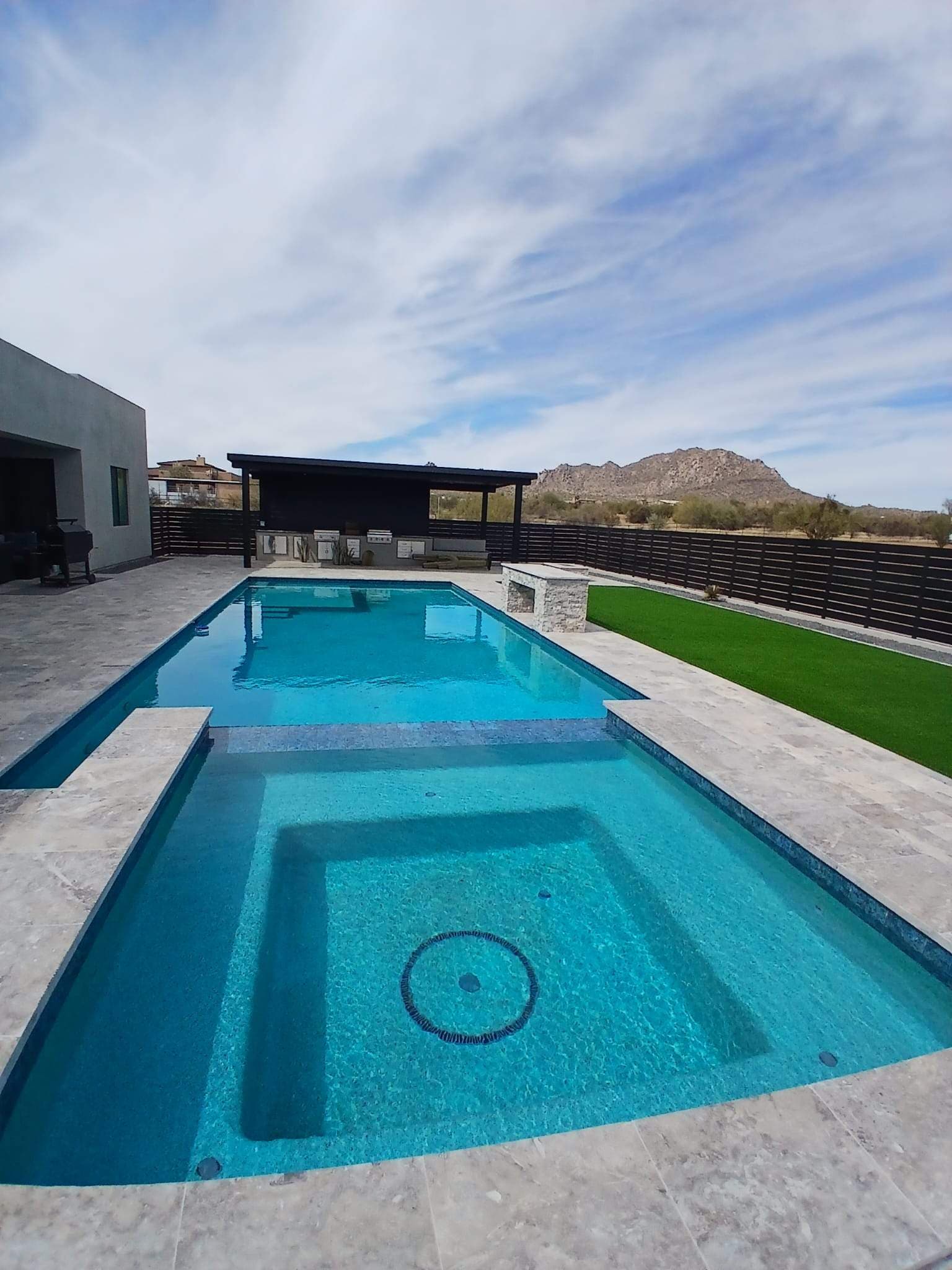 View from the near edge of a raised spa spilling into a long pool, with silver travertine decking, turf, a covered outdoor kitchen, and a desert mountain beyond