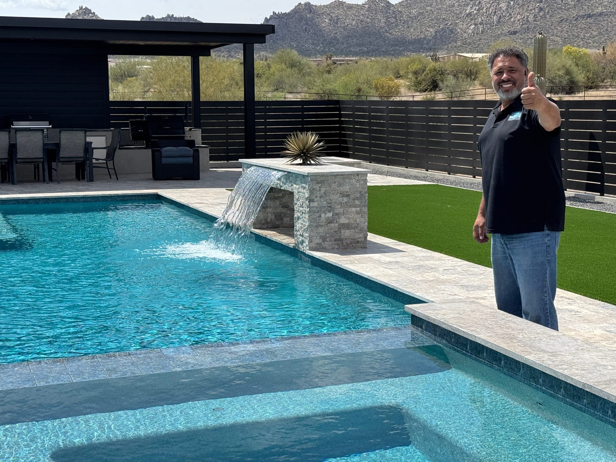 Waterfall pouring from a stacked-stone feature into the pool, with the covered outdoor kitchen and dining area behind