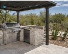 Stacked-stone outdoor kitchen with built-in grill under a steel pergola at sunset