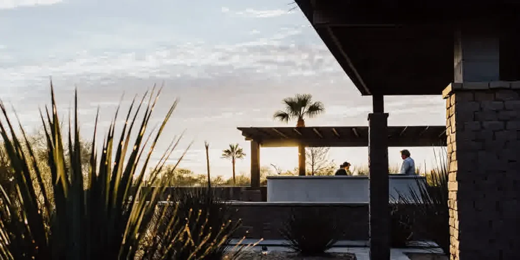 Luxury backyard transformation in Scottsdale Arizona with custom swimming pool, outdoor kitchen with stone countertops, cedar pergola, travertine decking, and desert landscaping at golden hour