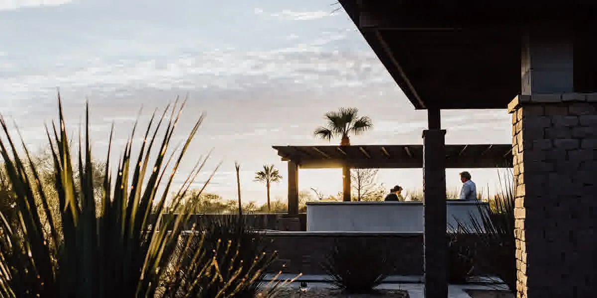 Luxury backyard transformation in Scottsdale Arizona with custom swimming pool, outdoor kitchen with stone countertops, cedar pergola, travertine decking, and desert landscaping at golden hour